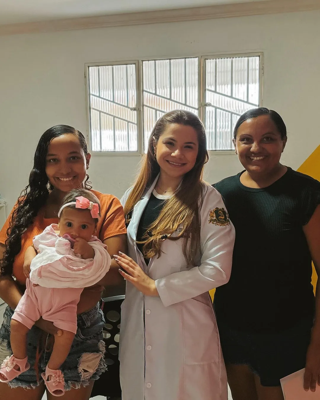 Mãe com a bebê no colo, a médica e outra participante sorriem juntas