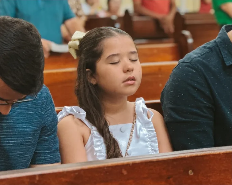 A girl with her eyes closed in prayer, seated among adults on a church pew